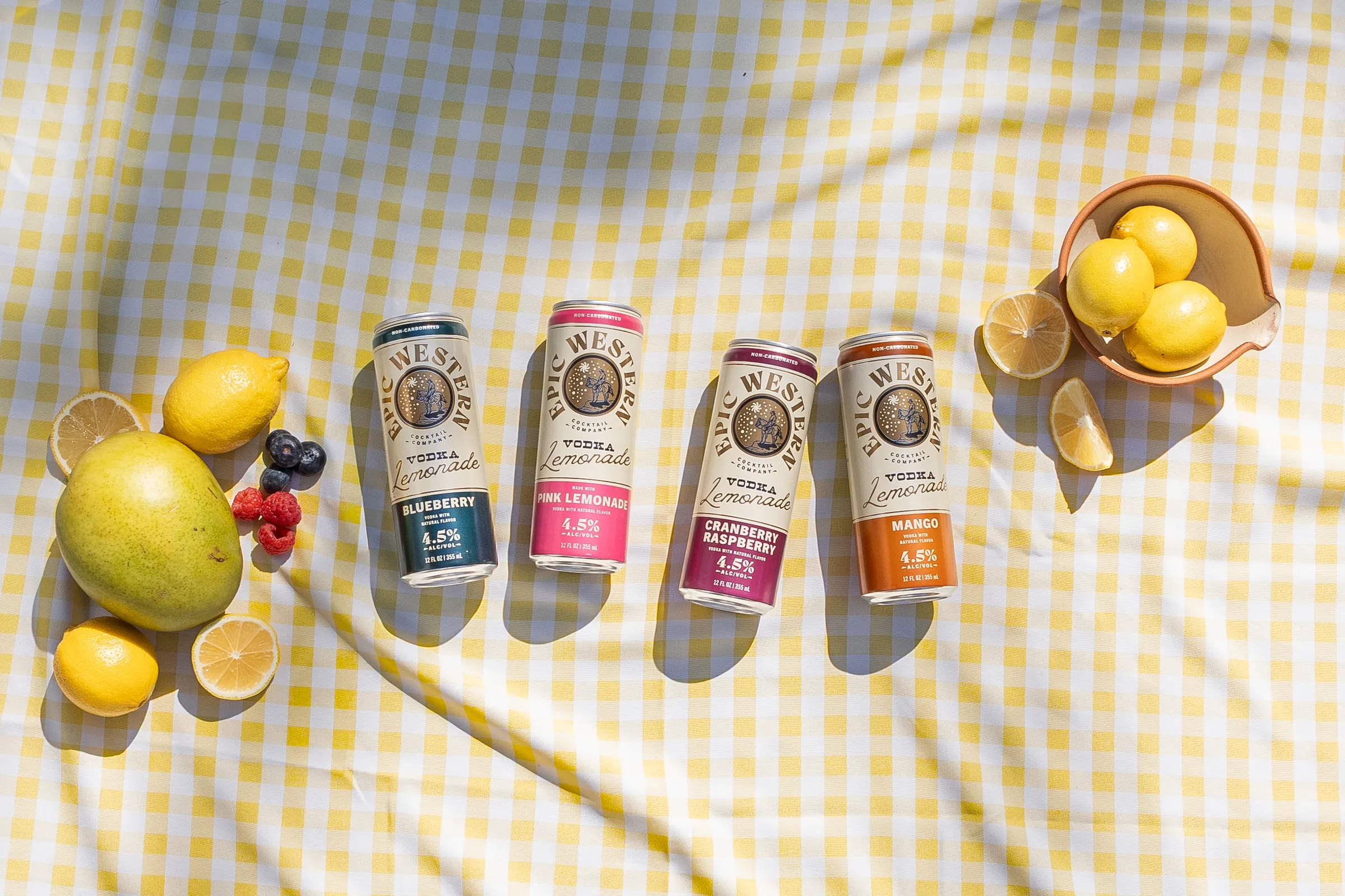 Four cans of Epic Western Vodka Lemonade on a yellow gingham picnic blanket with lemons and fresh fruit